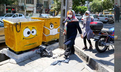 Iranian women walk past street bins, painted with a drawing of a mask and a sad face in a campaign t